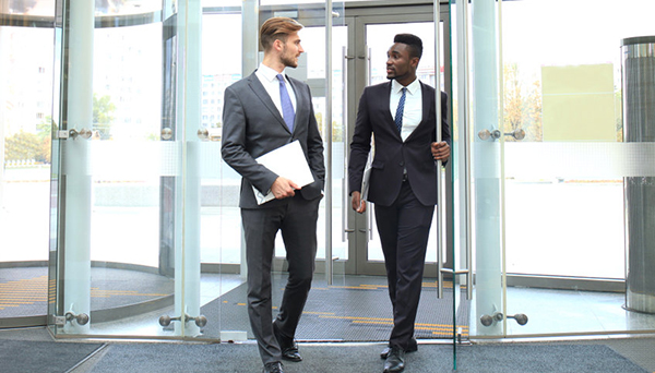Two multinational young businessmen entering in office building with glass doors.